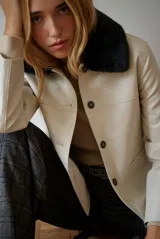 Chaqueta corta color beige con cuello de piel sintética negra, cierre frontal de botones y bolsillos laterales.