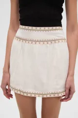 Falda corta blanca de algodón con pliegues y bordados geométricos en tonos rosados y verdes en el ruedo y la cintura.