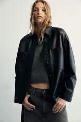 Camisa de efecto piel en color negro, con cuello solapa, manga larga y bolsillos de plastrón en el delantero. Presenta cierre frontal con botones ocultos por tapeta.