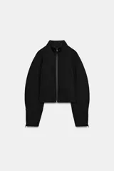 Chaqueta negra de cuello subido con cierre frontal de cremallera termosellada, manga larga con puños anchos y cremalleras laterales en los puños.
