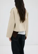 Chaqueta crop estilo gabardina corta color beige, de diseño recto, cuello alto tipo embudo, manga larga, bolsillos laterales y cierre cruzado de doble botonadura con botones oscuros. Con forro interior de poliéster.