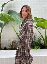 Camisa de corte holgado con estampado de cuadros en tonos marrones y beige. Presenta cuello clásico, cierre frontal con botones y mangas largas con aberturas laterales que permiten anudar la prenda en la cintura.