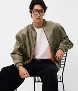 Campera bomber de poliuretano color verde militar con acabado efecto cuero, forro interior naranja, cierre frontal con cremallera, bolsillos delanteros con solapa, bolsillo utilitario en la manga y puños y ruedo elásticos.