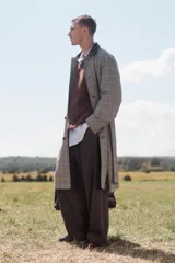 Trench de corte relajado con estampado de cuadros en tonos beige y marrón, cuello solapa en piel marrón, manga larga, bolsillos de vivo en cadera, cinturón ajustable y cierre frontal con botones.