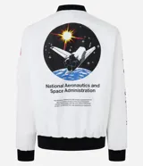 Campera tipo bomber blanca con detalles en negro, inspirada en uniformes espaciales de la NASA. Presenta parches con el logo de la NASA, la bandera de EE. UU. y texto en la manga que dice "Kennedy Space Center".