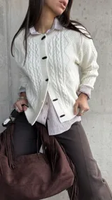 Cardigan de punto trenzado color natural, con cuello redondo, cierre frontal de botones negros y mangas largas con puños acanalados.