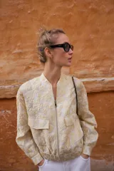 Chaqueta tipo bomber de cuello redondo y manga larga, confeccionada en tejido jacquard con diseño floral en tonos dorados y crema. Presenta cierre frontal con cremallera, bolsillos delanteros de plastrón con solapa y puños y bajo elásticos.