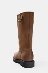 Bota de piel color marrón de caña alta, con detalle de hebilla metálica en la parte superior, efecto desgastado y suela dentada negra.