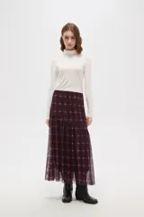 Falda larga de corte evasé con estampado de cuadros escoceses en tonos bordó y negro. Presenta un diseño de doble capa con una base opaca y una sobrecapa de tul semitransparente, con un corte asimétrico en la parte inferior.