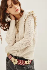 Cardigan de punto color beige con textura calada, cuello redondo y detalle de volados en los hombros. Presenta cierre frontal y puños acanalados.