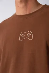 Remera de corte oversize color marrón, con cuello redondo y mangas cortas con hombros caídos. Presenta un bordado minimalista de un joystick en el centro del pecho y una etiqueta aplicada en la parte superior de la espalda.