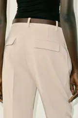 Pantalón tobillero de tiro medio, color beige, con pinzas delanteras y bolsillos frontales. Incluye un cinturón fino de cuero marrón con hebilla metálica dorada y cierre frontal con cremallera, botón interior y gancho.