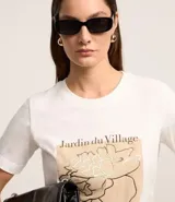 Remera blanca de algodón con estampado rectangular frontal con el texto "Jardin du Village" y un dibujo de una flor.
