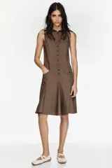 Vestido midi camisero color marrón, sin mangas, con cuello solapa y cierre frontal de botones. Presenta bolsillos delanteros con solapa y diseño con pliegues en el bajo.
