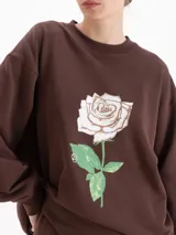 Buzo oversize color marrón de cuello redondo y manga larga, con estampa frontal de una rosa blanca y logo bordado en la espalda.