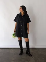 Vestido camisero corto negro de denim, con cuello clásico, cierre frontal con botones, mangas cortas, bolsillos cuadrados en el pecho y corte recto.