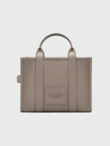 Cartera tipo tote de cuero granulado color tostado, con diseño estructurado y rectangular. Presenta doble asa superior fija y una correa larga ajustable y extraíble. En el frente, destaca el logo estampado con la inscripción 'THE TOTE BAG MARC JACOBS'.