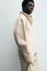 Cazadora de algodón color beige claro, corte relajado, con cuello subido y manga larga. Cierre frontal con cremallera oculta por solapa con cierres metálicos tipo alamar.