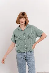 Camisa verde menta de tela arrugada, con cuello clásico, botones frontales, mangas cortas y dos bolsillos delanteros con pespuntes. La base es levemente elastizada y se anuda en el frente.