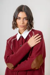 Saco de punto color beige, con cierre frontal de botones marrones y detalle de costuras decorativas oscuras en los hombros. Presenta un diseño clásico con cuello redondo.