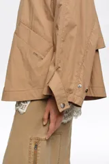 Gabardina corta de línea A y tejido técnico color tostado, con cuello alto, cierre frontal con cremallera metálica, mangas largas con trabillas ajustables y bolsillos de vivo.