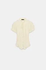 Top color crema con cuello redondo y lazada, escote en pico y mangas cortas abullonadas con elástico. Presenta un tejido elástico tipo nido de abeja y cierre frontal con botones.