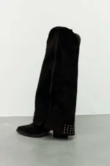 Bota de caña alta estilo slouchy confeccionada en gamuza sintética de color negro, con punta fina y taco bajo. Presenta solapa superior plegada con apliques de tachas metálicas plateadas y una tira con hebilla decorativa en la parte superior de la caña.