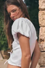 Top crop blanco confeccionado en mezcla de algodón, con escote en V, mangas corta abullonadas, detalles fruncidos y cierre frontal con dos lazos.