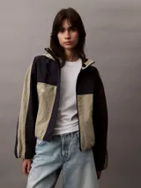 Campera corta con capucha, confeccionada en tejido ripstop de nailon en dos tonos: beige y negro. Tiene corte relaxed, cierre de cremallera y botón de presión, bolsillos con cremallera en la cintura, bajo con cordón elástico, forro de malla transpirable y logo CK bordado en el pecho.