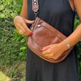 Cartera bandolera de eco-cuero marrón, de diseño redondeado con solapa frontal y bolsillo con cierre. Incluye correa ajustable y desmontable con patrón geométrico.