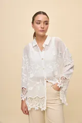Camisa blanca de calce relajado, con cuello clásico, mangas 3/4 y cierre frontal de botones. Presenta bordados florales en relieve y detalles de crochet calado en los puños y el ruedo.
