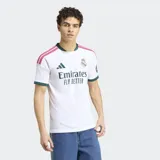 Camiseta de futbol Adidas del Real Madrid, primera equipacion. Diseño en color blanco con detalles en verde oscuro en el cuello y los puños, y franjas fucsias en los hombros. Cuenta con el logo de la marca, el escudo del club bordado y el patrocinador oficial en el frente.