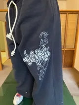 Pantalón de jogging negro de corte baggy, confeccionado en algodón. Presenta una estampa plastisol con bordado decorativo en la pierna izquierda, cintura elástica con cordón largo ajustable y botones en el ruedo para regular la botamanga.