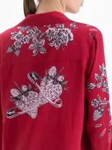 Camisa de manga larga color rojo, con cuello clásico y cierre frontal de botones. Presenta un estampado artístico que combina motivos florales en tonos rosados con ilustraciones de alfileres de gancho.