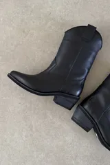 Bota texana de cuero color negro, de caña media, con taco cuadrado y puntera ligeramente perfilada. Presenta costuras decorativas y tirador superior.