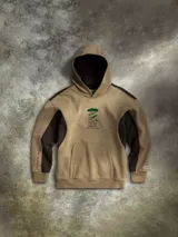 Buzo con capucha tipo canguro en color beige con paneles laterales en color marrón oscuro. Presenta un diseño bordado central de árboles y detalles gráficos en las mangas. Incluye bolsillo frontal tipo canguro, capucha forrada y puños acanalados.