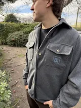 Camisa de lana gris melange de corte clásico y atemporal, con cuello camisero, bolsillos frontales con solapa y botones, y detalle bordado en el pecho.