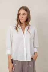 Camisa blanca de corte clásico con cuello camisero, cierre frontal de botones y mangas 3/4 con ligero fruncido en los hombros.