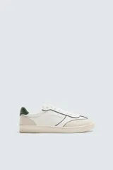 Zapatilla deportiva de hombre, estilo retro, con capellada blanca y detalles en verde oscuro en el talón y ribetes negros. Presenta perforaciones en el lateral y suela de goma color crema.
