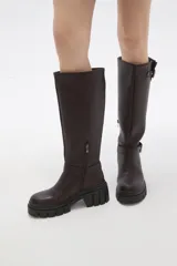 Bota de caña alta color negro, confeccionada en eco cuero, con suela track de goma y detalle de hebilla metálica en el tobillo y en la parte superior de la caña.