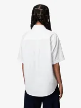 Camisa blanca de manga corta y corte holgado, confeccionada en popelina de algodón ligero. Presenta cuello clásico, cierre frontal abotonado y logo bordado en el pecho izquierdo.