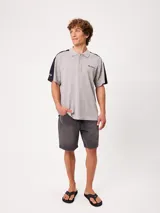 Remera polo gris melange de ajuste holgado, con cuello y cierre frontal de botones. Presenta detalles en azul marino y blanco en los hombros y mangas, y el logo de la marca bordado en el pecho.