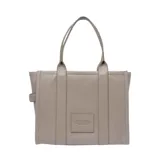 Bolso tipo tote de gran tamaño, confeccionado en cuero marrón con textura granulada. Presenta el logo de la marca estampado en el frente con la inscripción The Tote Bag Marc Jacobs. Cuenta con asas superiores fijas y un diseño estructurado y espacioso.