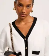 Cardigan de punto blanco con escote en V, botones metalizados, bolsillos falsos, cuello y vista contrastantes en negro, y bajo acanalado.