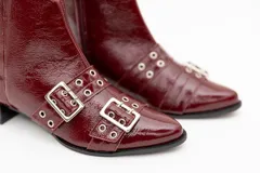 Botas cortas de cuero charol color negro, con aplique de hebillas con ojalillos y cierre lateral. Taco cuadrado bajo.