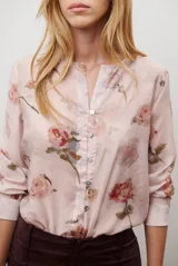 Camisa de manga larga con cuello redondo y cierre frontal de botones. Presenta un estampado floral en tonos azules y marrones sobre una base de color rosado claro.