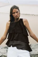 Top de cuello redondo y manga sisa, confeccionado en tejido con acabado satinado. Presenta volantes en cascada y un diseño de bajo asimétrico, con cierre en la espalda mediante pequeña abertura y botón.