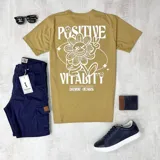 Remera color mostaza con estampa en la espalda que incluye una flor con una cara sonriente, la palabra "Positive" y la frase "Stepping Beyond Yourself Duxon Jeans".