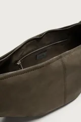 Bolso de hombro tipo hobo con diseño de media luna, confeccionado en gamuza color marrón. Cuenta con un asa larga fija de cuero negro y cierre superior con cremallera.