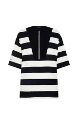 Blusa de punto con estampado de rayas horizontales en blanco y negro. Presenta un corte holgado, cuello redondo con panel frontal sólido en contraste, cierre de cremallera metálica, mangas cortas anchas y un parche bordado en la manga derecha.
